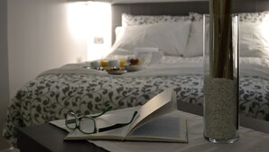 Un lit bien fait, recouvert d’une couvre-lit à motifs, une table de chevet avec un livre et des lunettes, et un grand vase en verre rempli de sable.