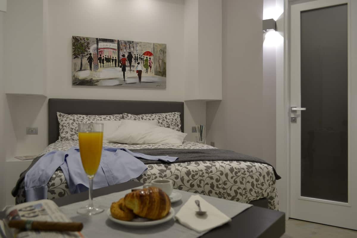 A hotel room with a bed, a bedside table with a croissant, a glass of orange juice, and a painting on the wall.
