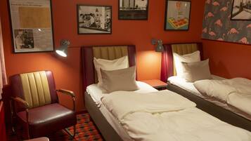 A hotel room with two beds, a chair, and a table. There are framed pictures on the wall.
