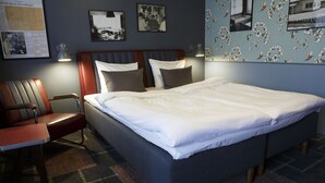 A neatly made bed with white linens, a wooden headboard, and a plaid-patterned rug. There is a single chair with a wooden frame and a cushion. The room has a dark blue wall with floral wallpaper and a framed picture above the bed. - Wittrup Motel (Albertslund)