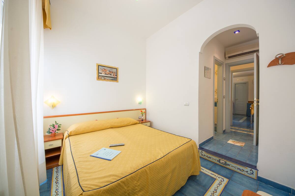 A hotel room with a bed, bedside table, lamp, and a small potted plant.