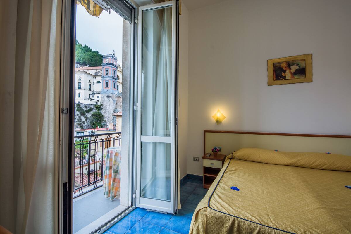 A hotel room with a bed, a nightstand, a wall-mounted lamp, and a balcony with a view of buildings.