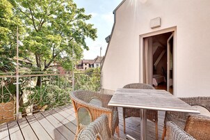 Un balcón con mobiliario de mimbre y una mesa, con vistas a un jardín y edificios cercanos.