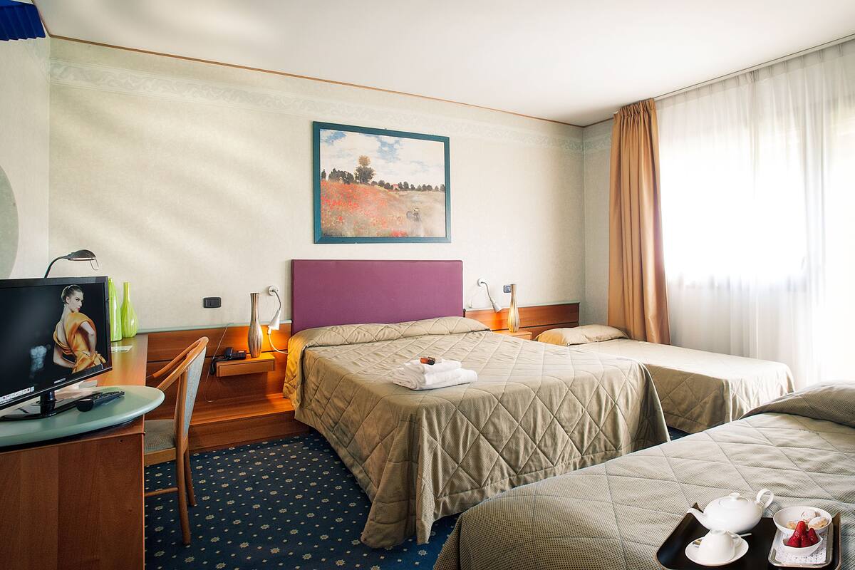 Une chambre d’hôtel avec deux lits, un bureau avec un ordinateur, un tableau au mur et une fenêtre avec des rideaux.