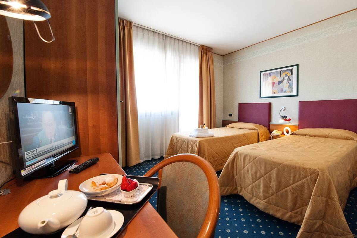 Une chambre d’hôtel avec deux lits, une télévision, un bureau avec une théière et une assiette de nourriture, et un tableau accroché au mur.