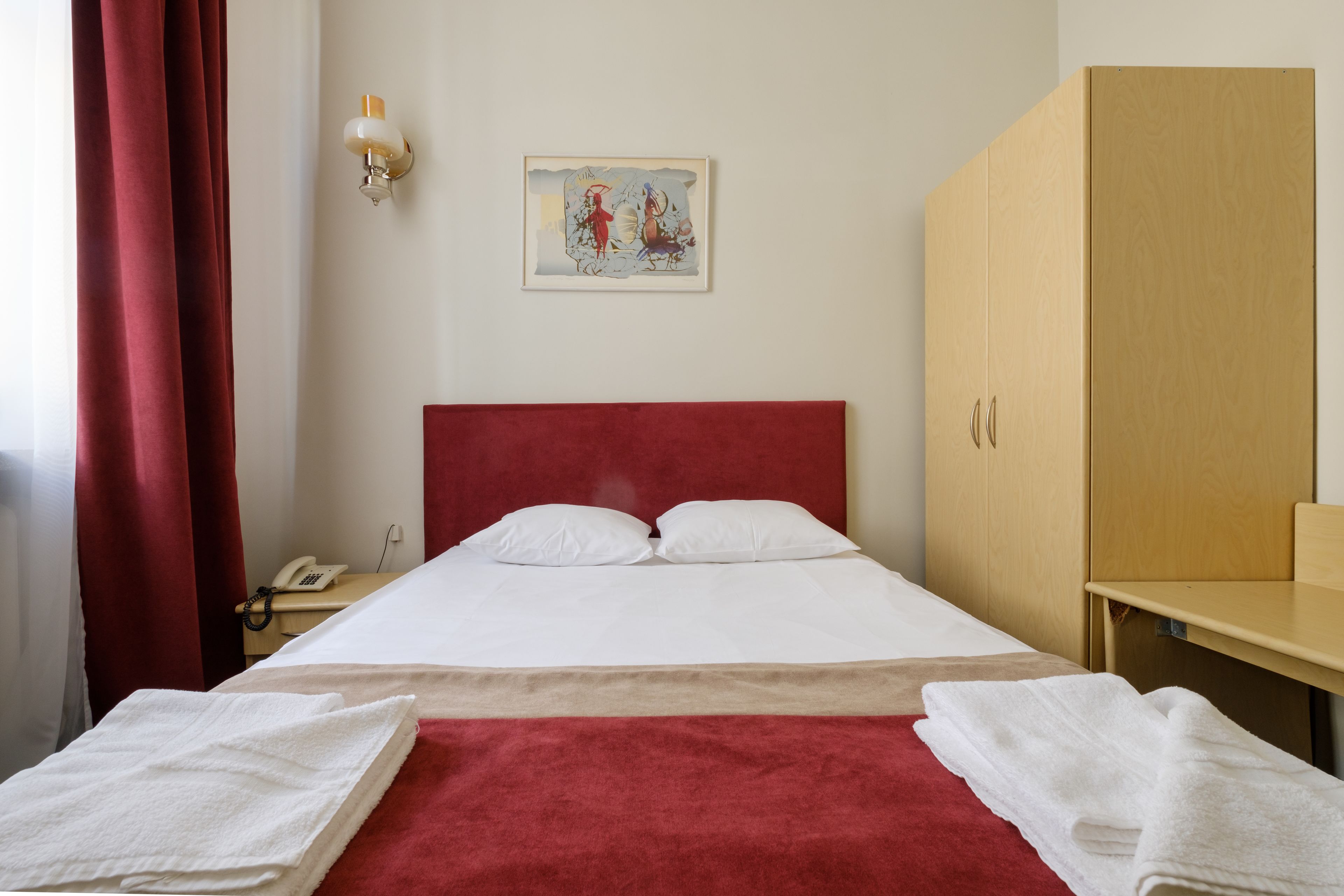 Une chambre d’hôtel avec une tête de lit rouge, du linge de lit blanc, un plaid rouge et une armoire en bois.