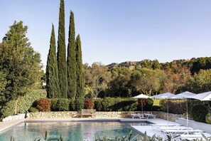 A pool area with lounge chairs and umbrellas, surrounded by tall trees and a stone wall. - Du Côté des Olivades (Paradou)