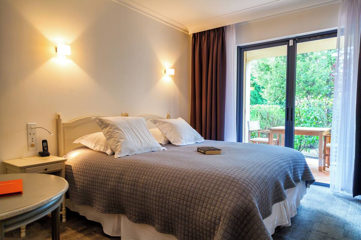 A hotel room with a large bed, bedside tables, a small round table, and a view of the outdoors through a sliding glass door.