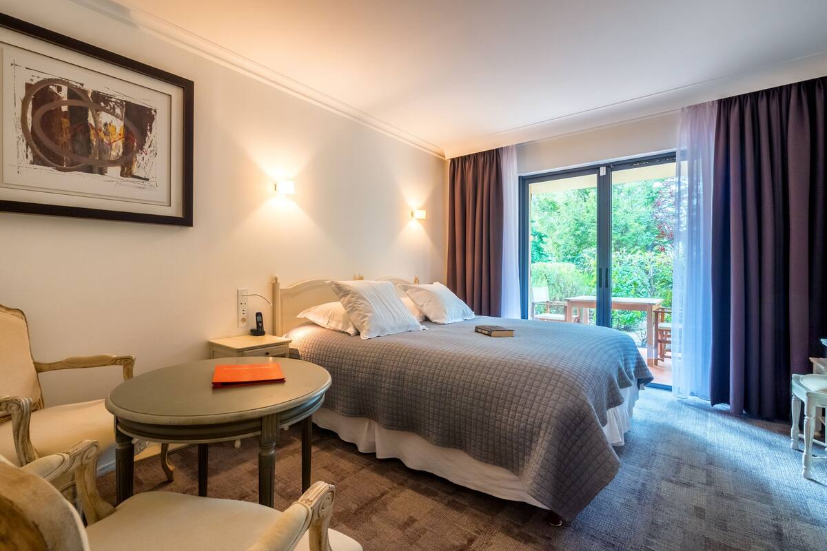 A hotel room with a bed, a round table, a chair, a framed picture, and a view of the outdoors.
