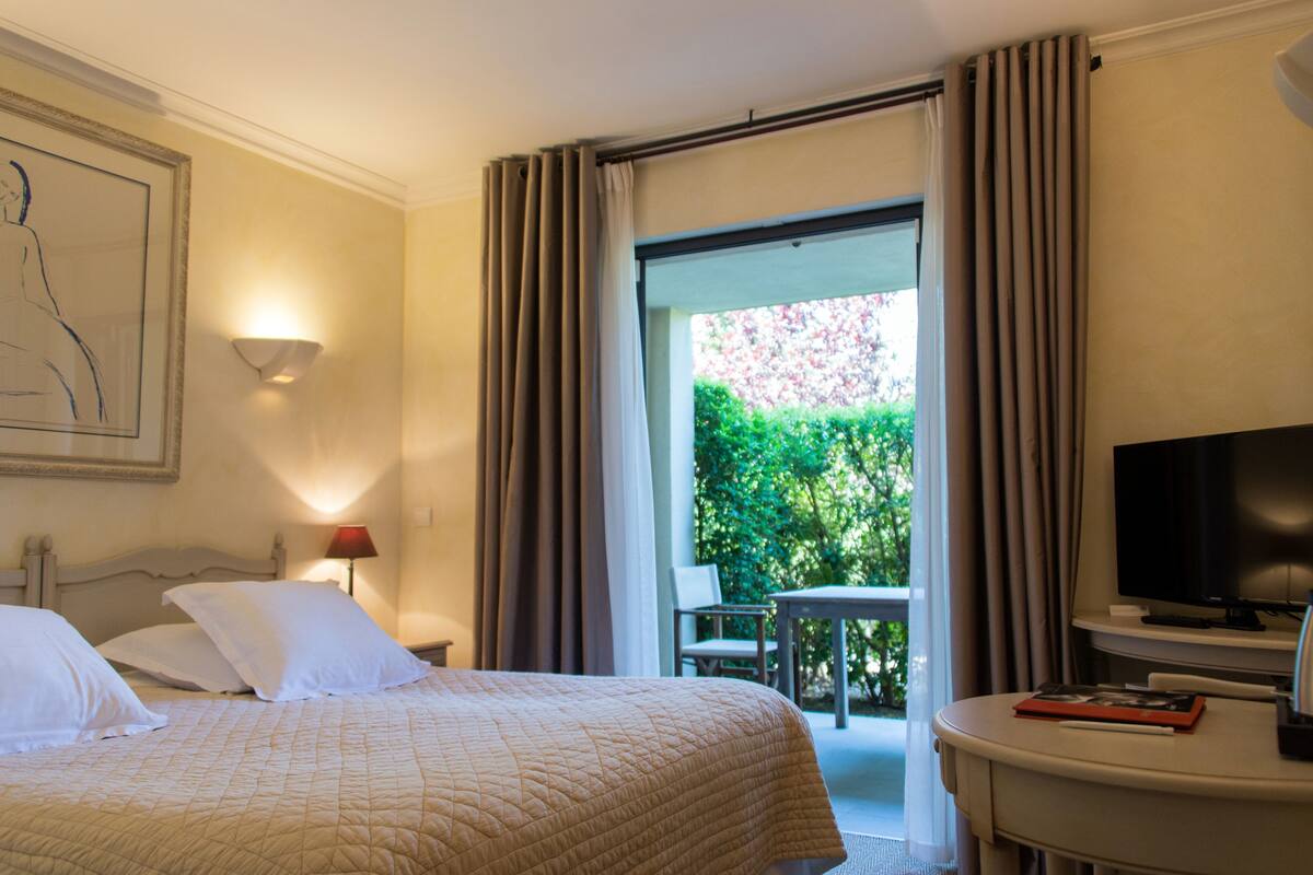 A hotel room with a bed, a television, a desk, and a view of a garden through a sliding door.