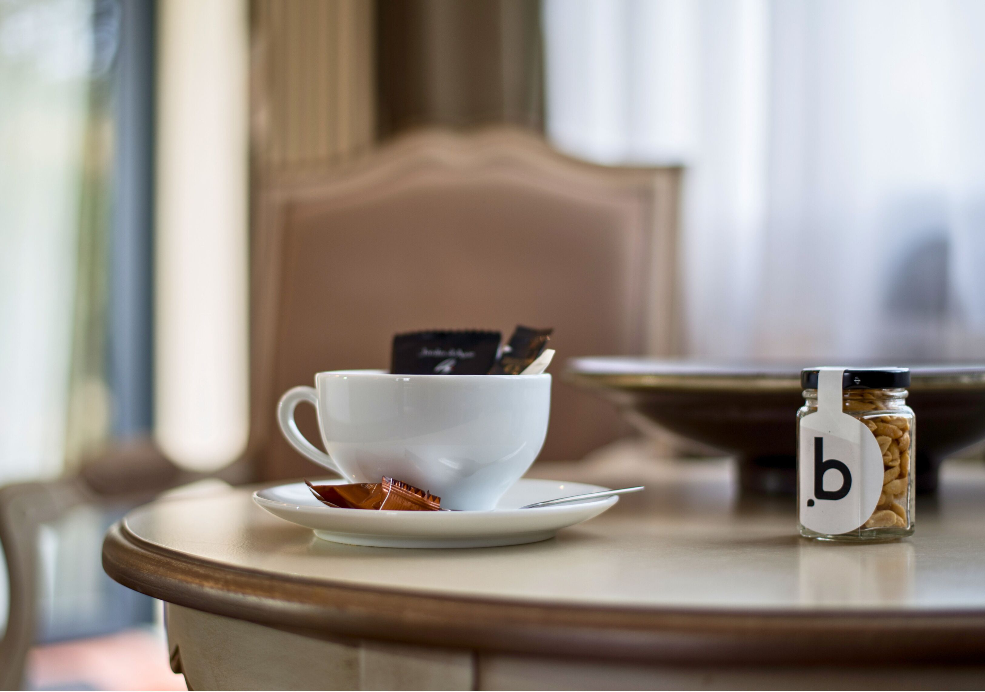 A white cup with a tea bag on a saucer, a jar labeled 'b' with coffee beans, and a wooden chair.