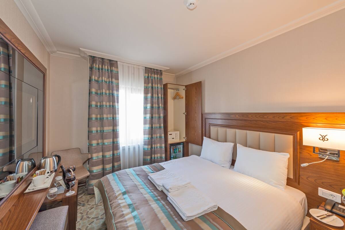 A hotel room with a neatly made bed, a desk with a telephone and a cup, a mirror, and a wardrobe.