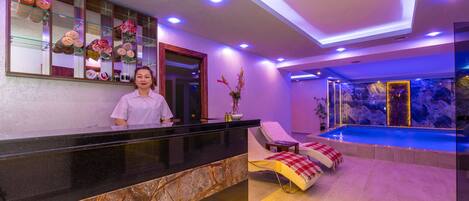 A reception area with a marble counter, a reception desk, a lounge chair, and a pool area with a view of the pool.