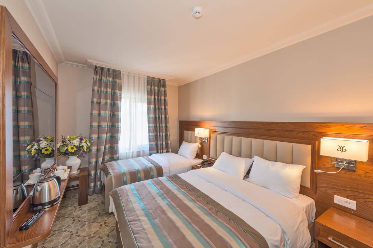 A hotel room with two beds, a wooden headboard, a nightstand with a lamp, a mirror, a dresser, and a window with curtains.