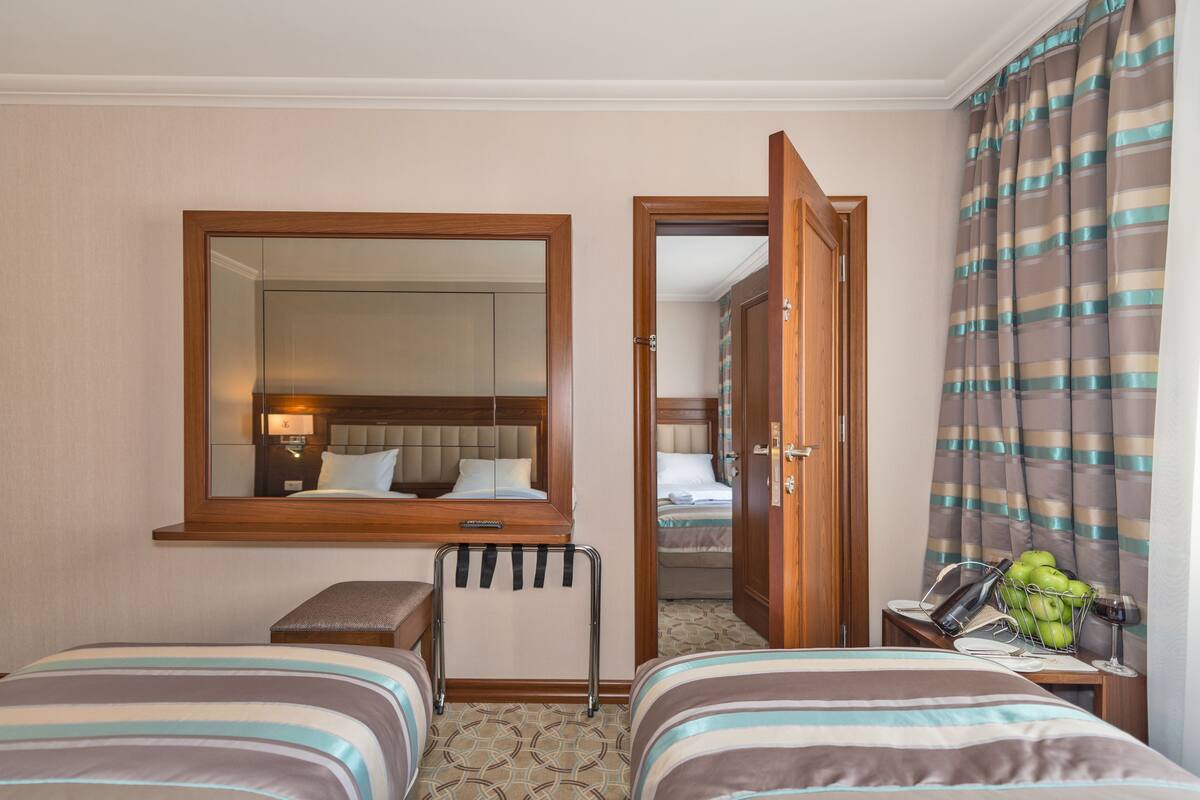 A hotel room with two beds, a bedside table, a mirror, a window with curtains, and a door leading to another room.