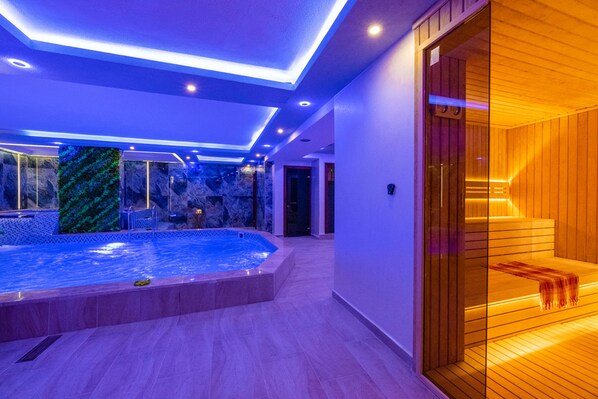 A modern indoor swimming pool with a glass enclosure, wooden sauna area, and a stone wall with a green pattern.