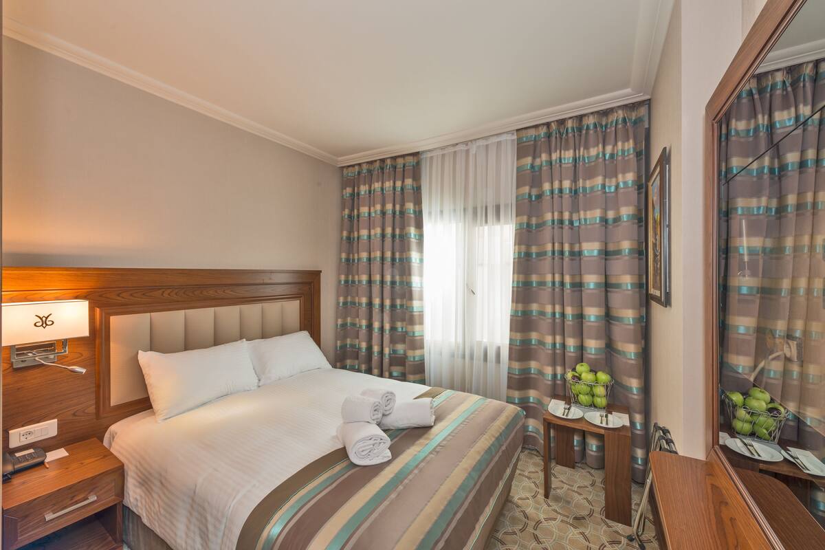A hotel room with a neatly made bed, a bedside table with a lamp, a small table with a fruit basket, and a mirror.