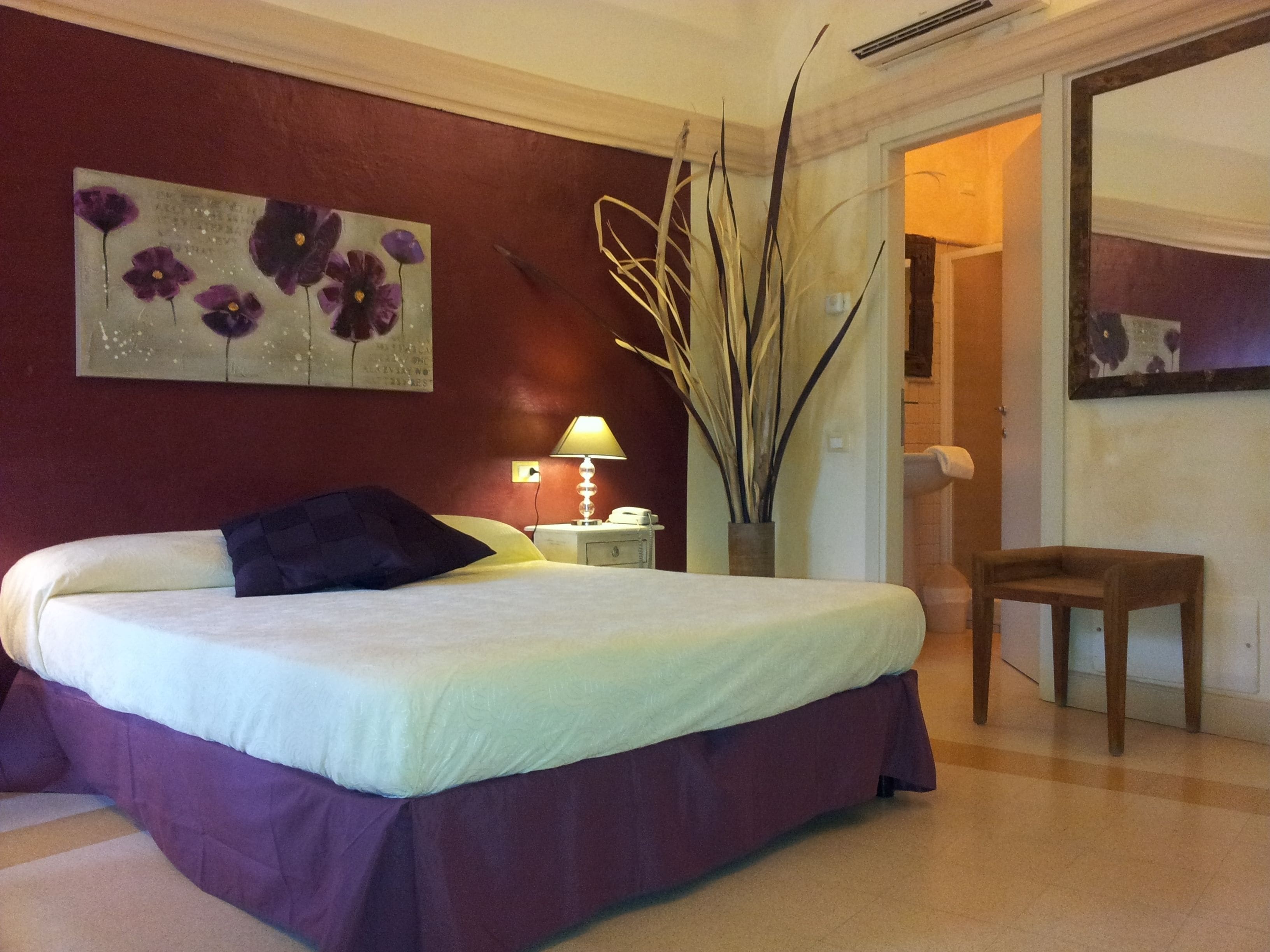 A bedroom with a bed, a nightstand, a lamp, a vase with tall plants, and a chair.