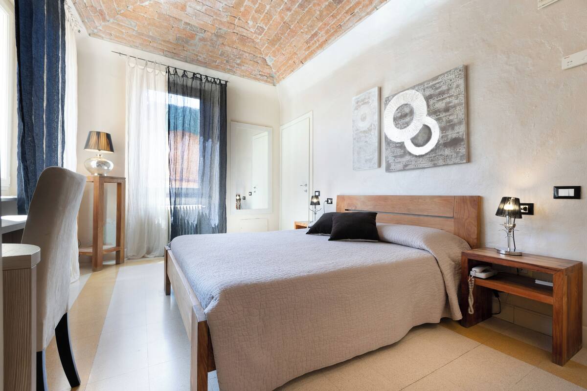 A modern bedroom with a large bed, a wooden nightstand, a desk with a lamp, and a chair.