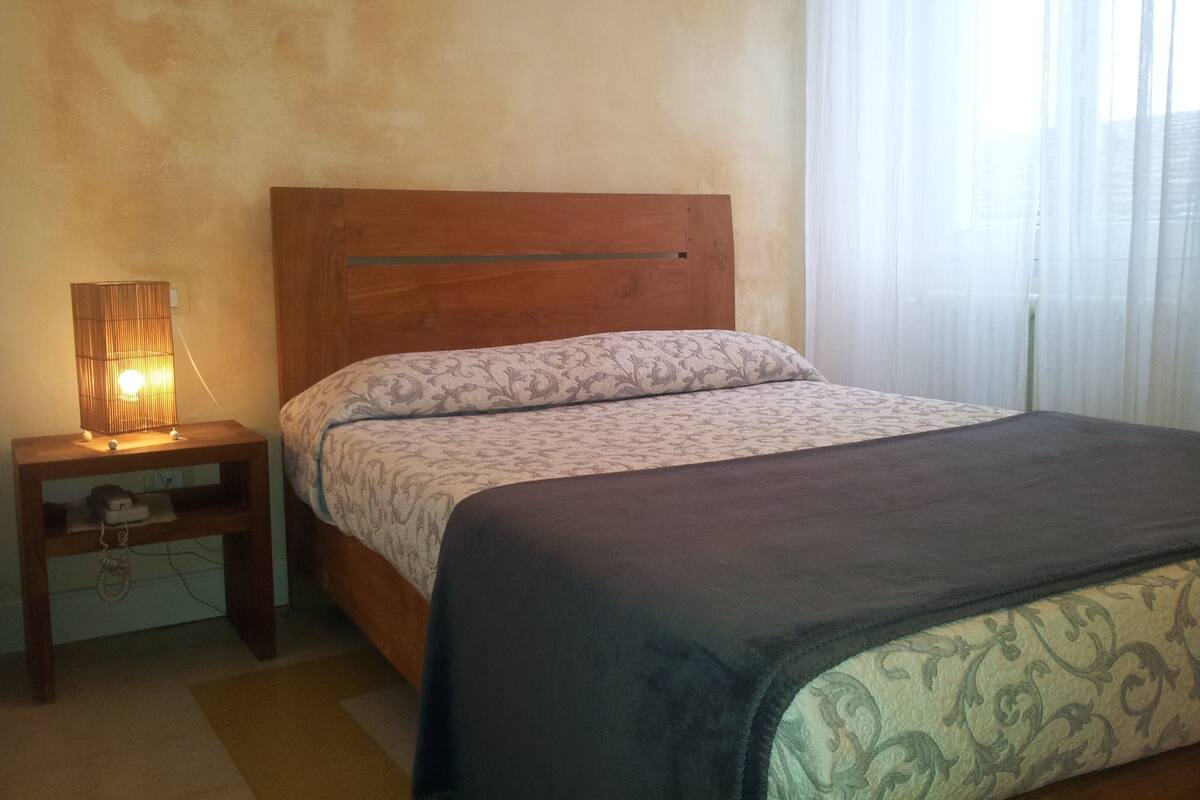 A bedroom with a wooden headboard, a bedside table with a lamp, a patterned bedspread, and a window with sheer curtains.