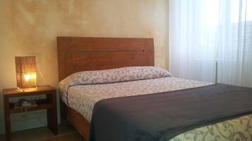 A bedroom with a wooden headboard, a bedside table with a lamp, a patterned bedspread, and a window with sheer curtains.