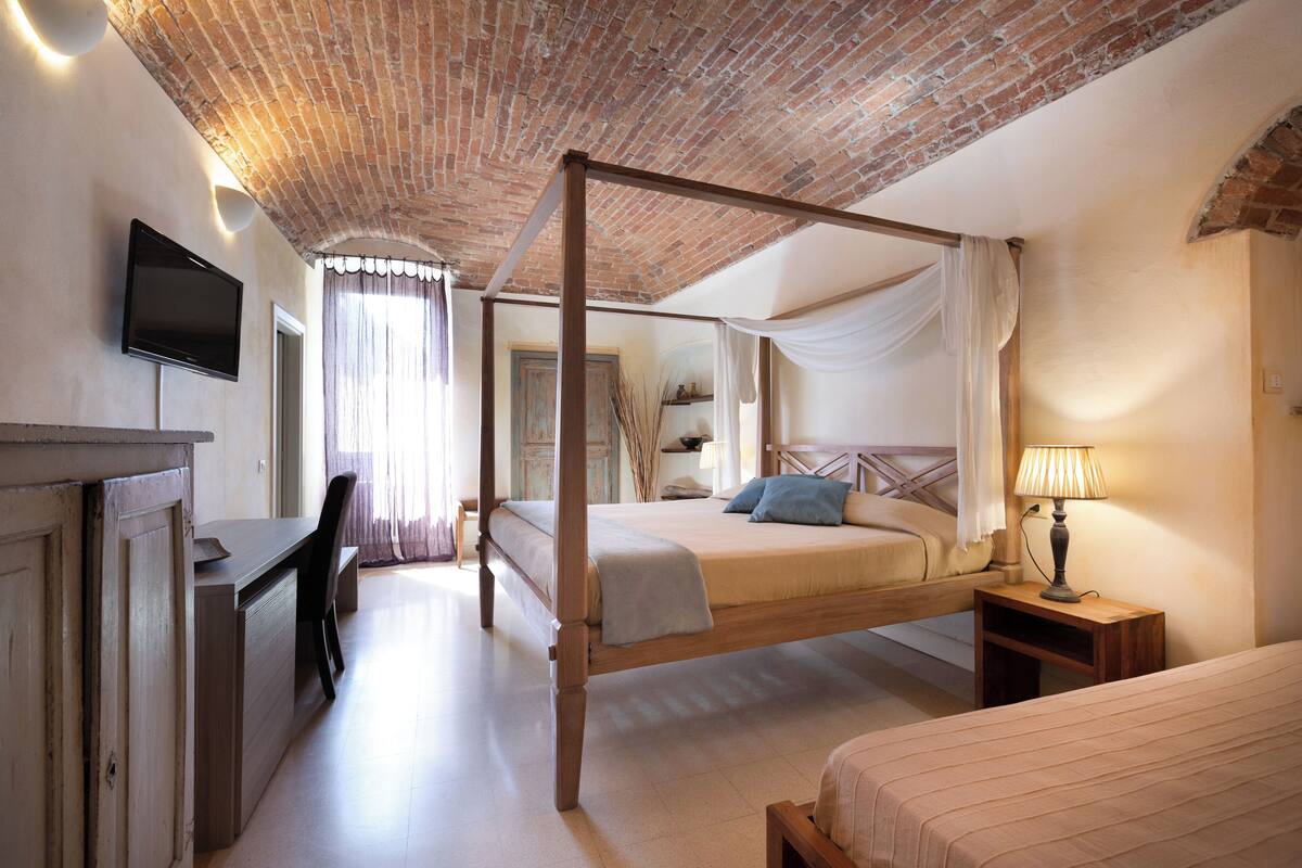 A four-poster bed with a wooden frame, a desk with a chair, a television mounted on the wall, and a bedside table with a lamp.