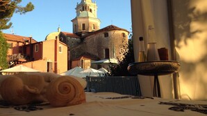 A table with a patterned tablecloth, a rolled-up towel, and a candle holder with candles. In the background, there is a building with a bell tower and a flag. - Hotel Relais al Convento (Vezzano Ligure)
