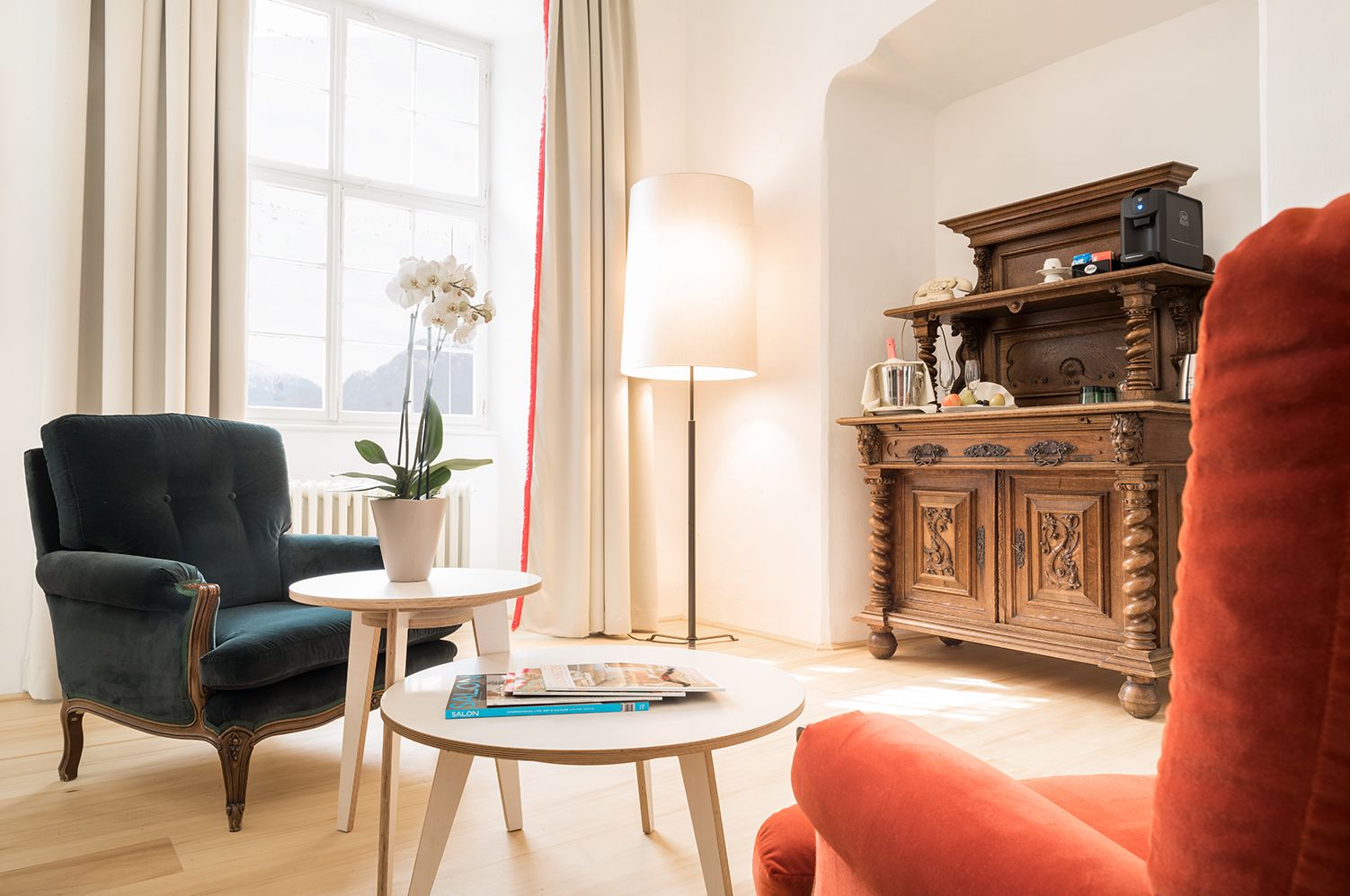 A living room with a dark green armchair, a round wooden table with a magazine, a floor lamp, and a wooden cabinet with decorative items.
