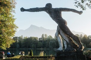 Una estatua de un hombre con los brazos extendidos en un parque con un lago y montañas al fondo.