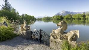 A serene lakeside setting with stone statues of horses and a metal gate, surrounded by lush greenery and mountains in the background. - Schloss Leopoldskron (Salzburg)