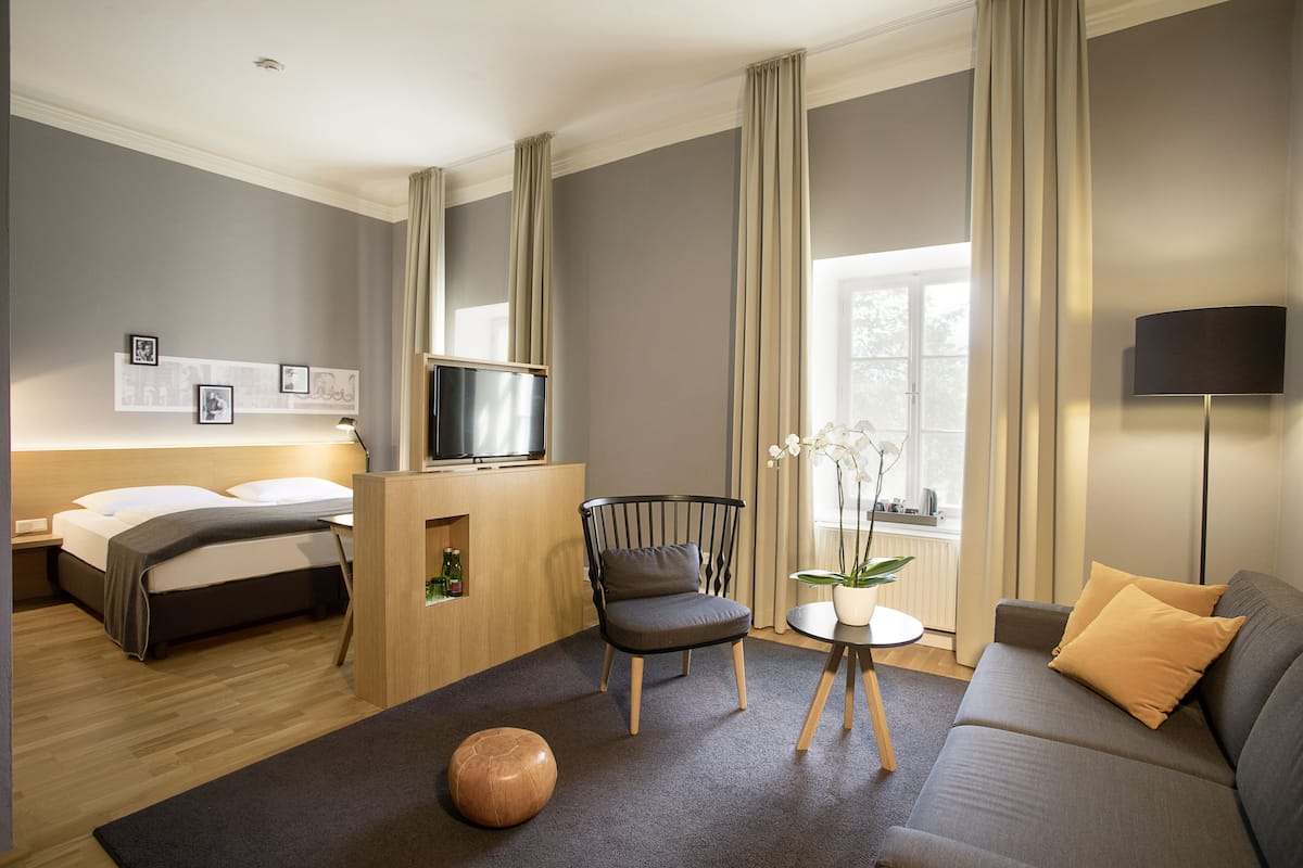 Quarto de hotel moderno com cama, sofá, poltrona, mesinha e televisão.