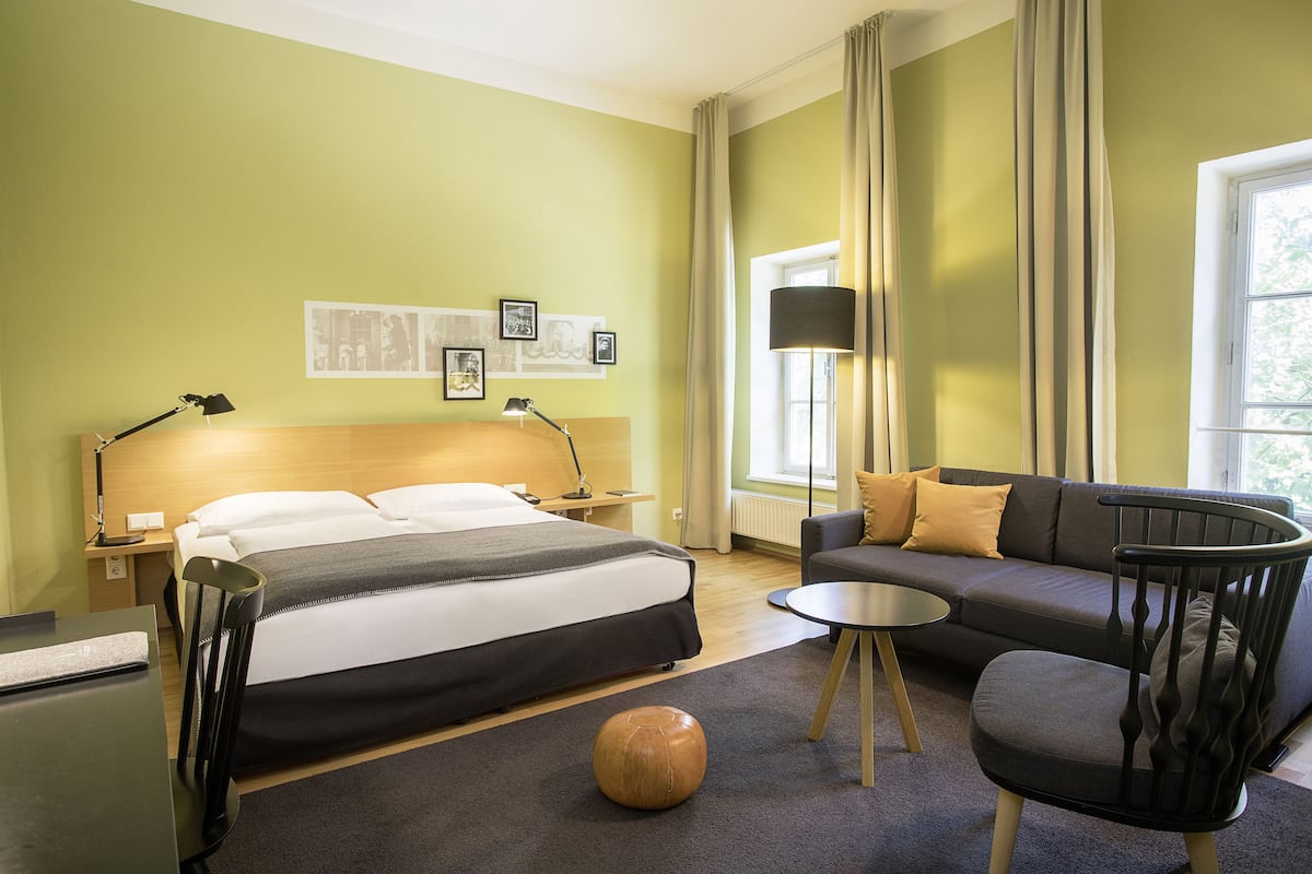 Quarto de hotel com cama, sofá, mesa de centro redonda e um banquinho de madeira.