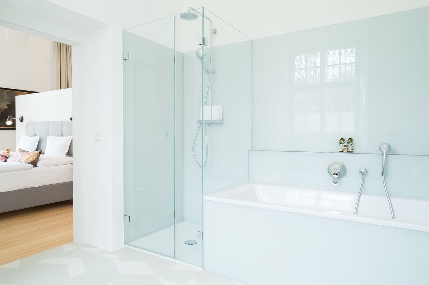 Un baño moderno con ducha envuelta en vidrio, una bañera blanca y una mesita de noche, con una cama en la habitación contigua.