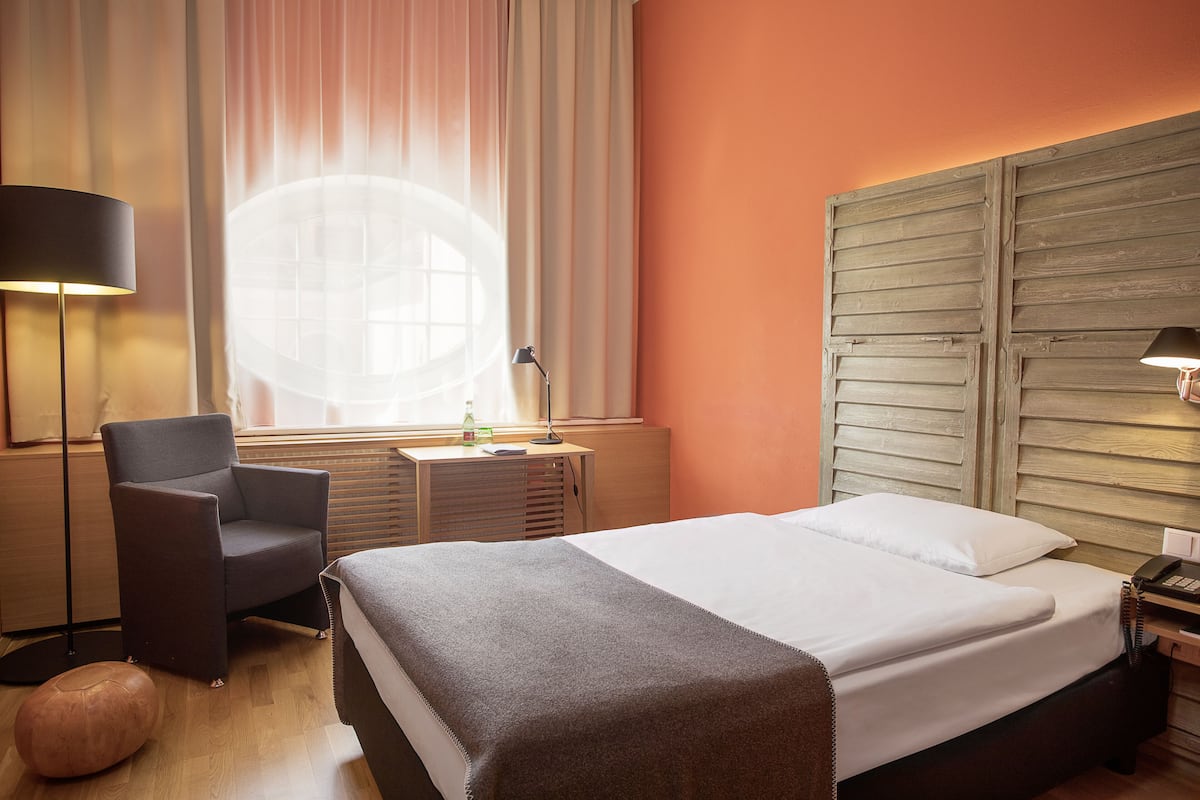 Quarto de hotel com cama, uma cadeira, escrivaninha, luminária e janela com cortinas.