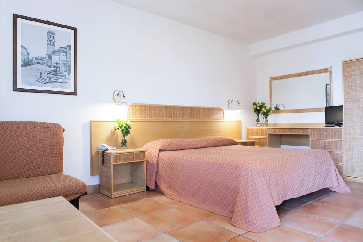 A hotel room with a bed, a desk, a chair, a nightstand, a mirror, and a framed picture on the wall.