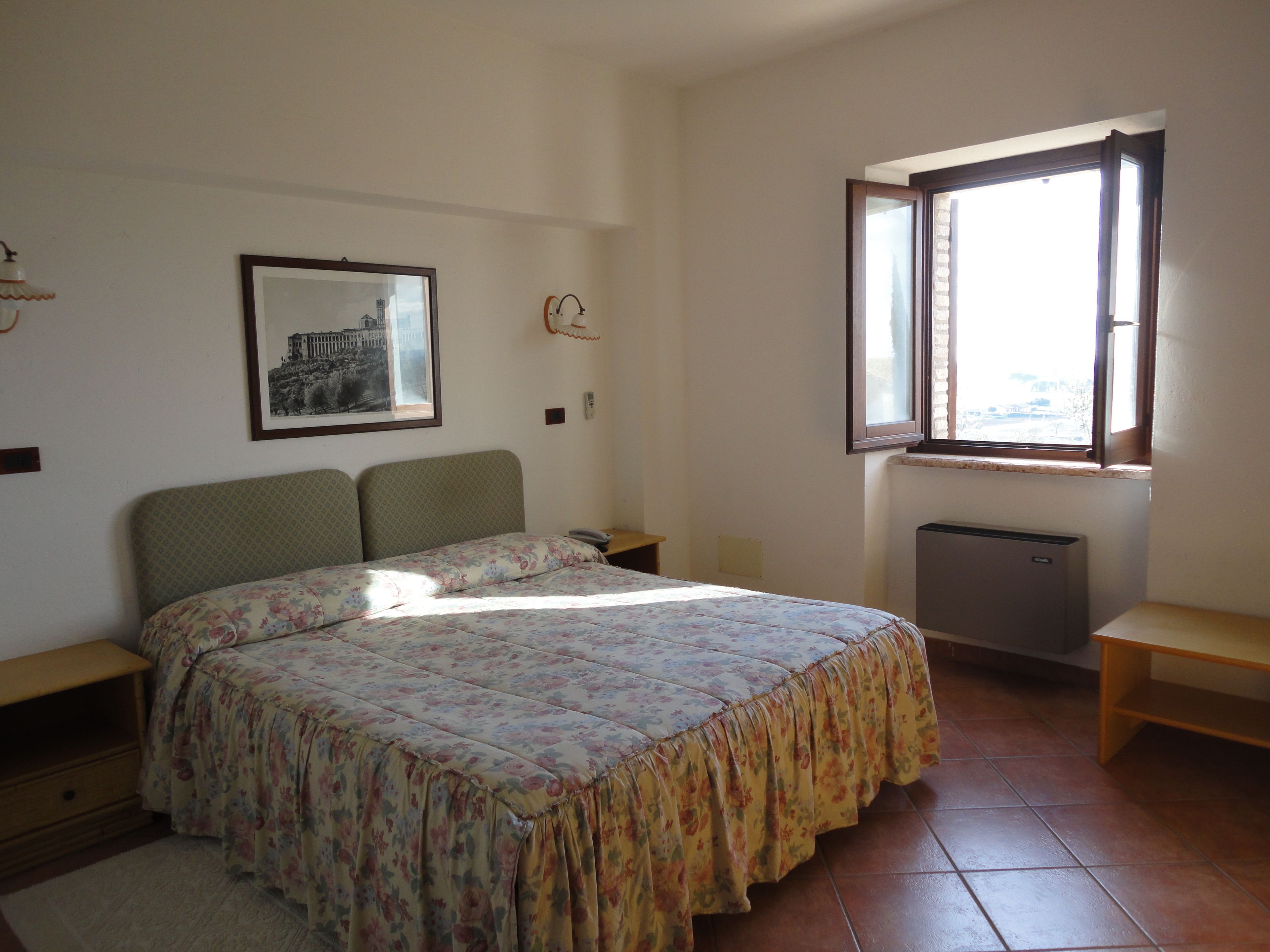 A bedroom with a bed, a nightstand, a bedside table, a radiator, a window, and a framed picture on the wall.