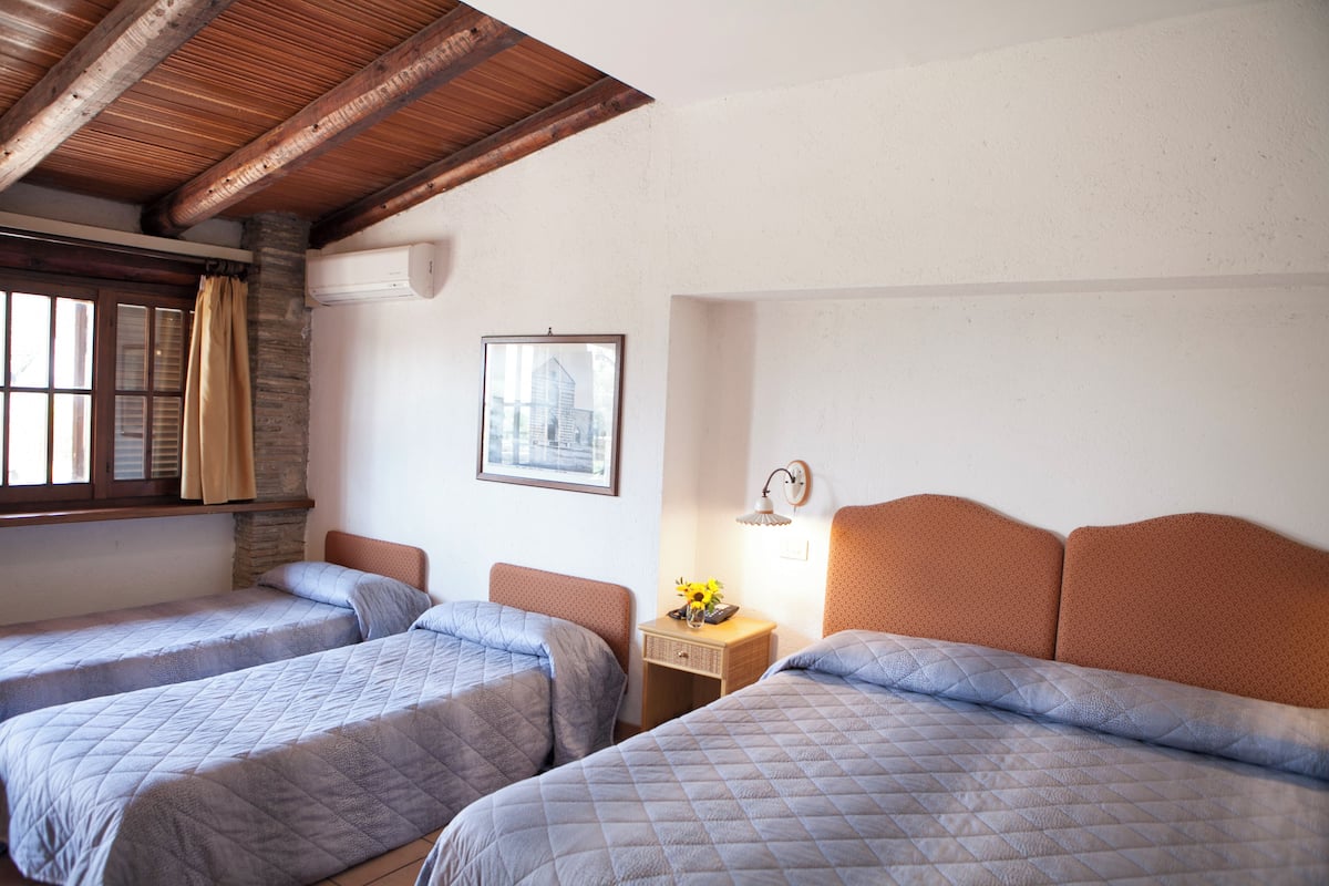 A room with two beds, a window with curtains, a wall-mounted air conditioner, a framed picture, and a bedside table with a lamp.