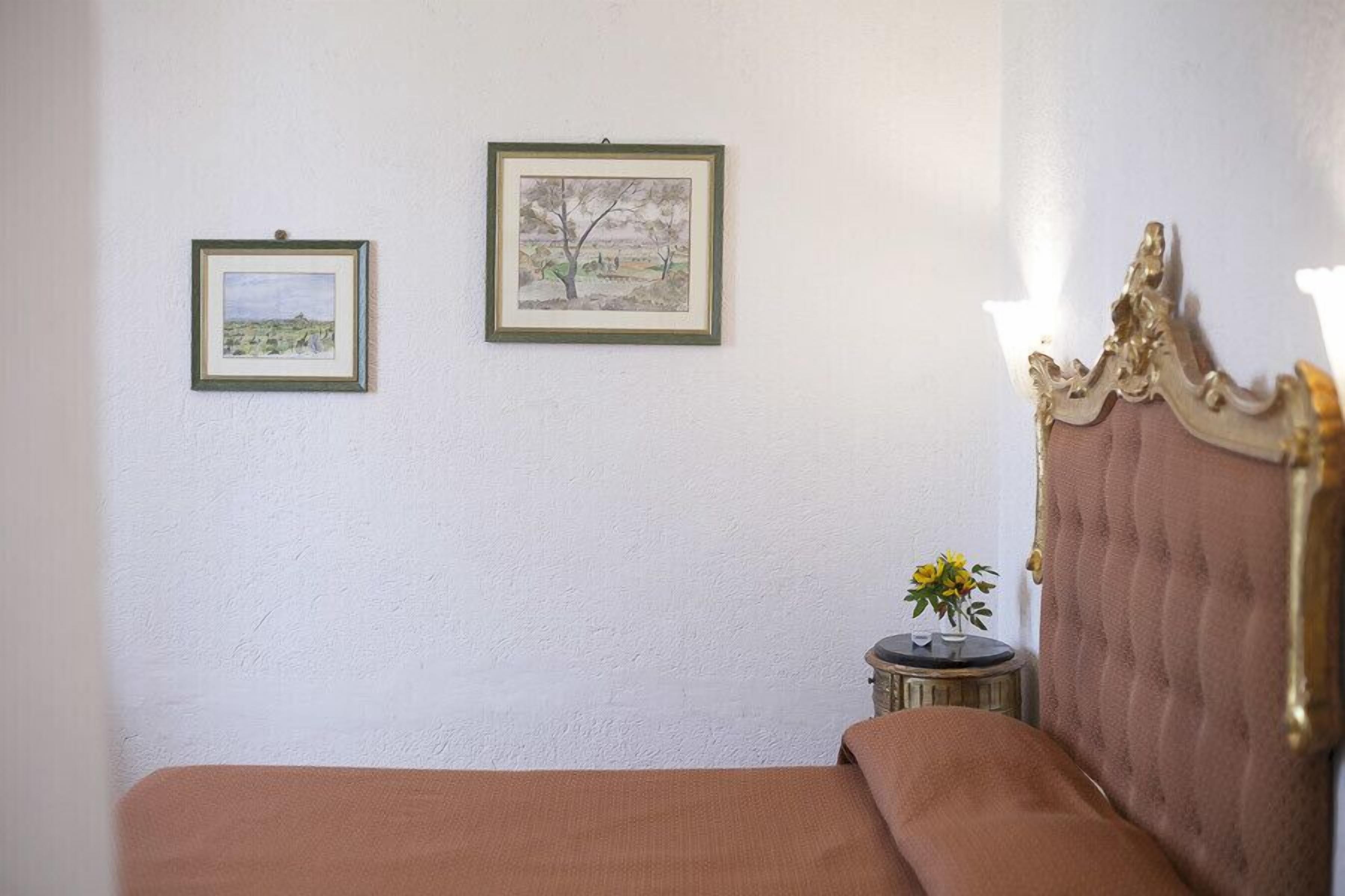 Un dormitorio con una cama, dos cuadros en la pared, una mesita redonda con un florero y un cabecero decorativo.
