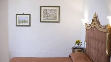 Un dormitorio con una cama, dos cuadros en la pared, una mesita redonda con un florero y un cabecero decorativo.