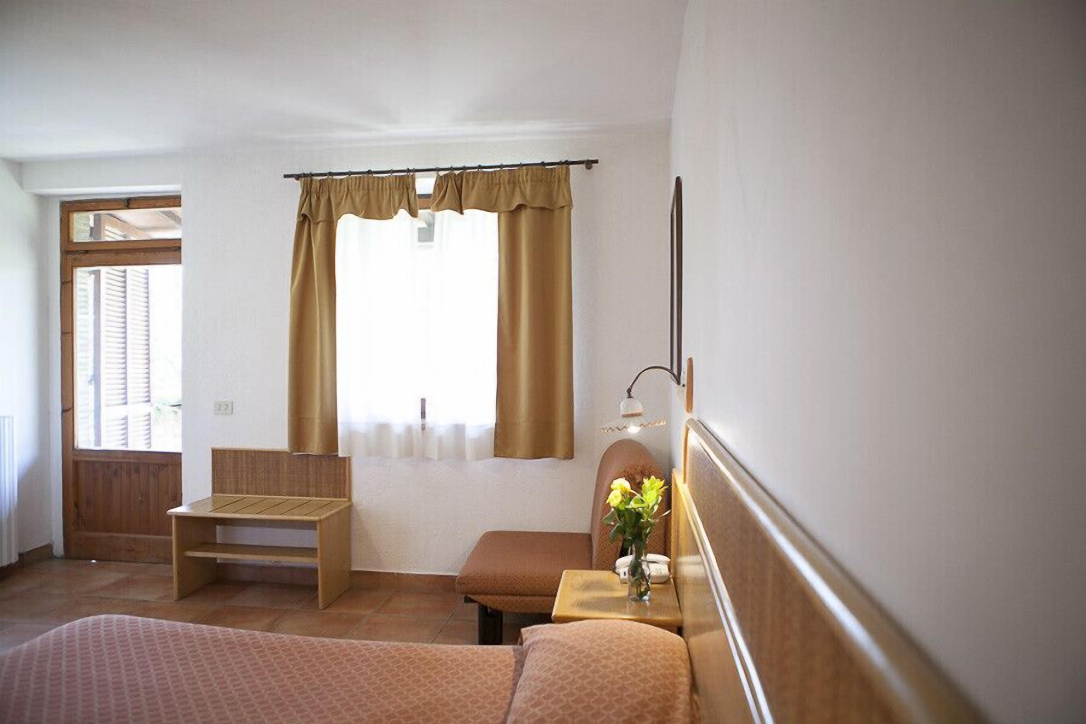 A hotel room with two beds, a wooden bench, a small table with a vase of flowers, and a window with curtains.