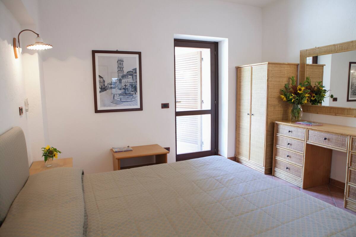 A bedroom with a bed, a desk, a chair, a wardrobe, a mirror, and a framed picture on the wall.