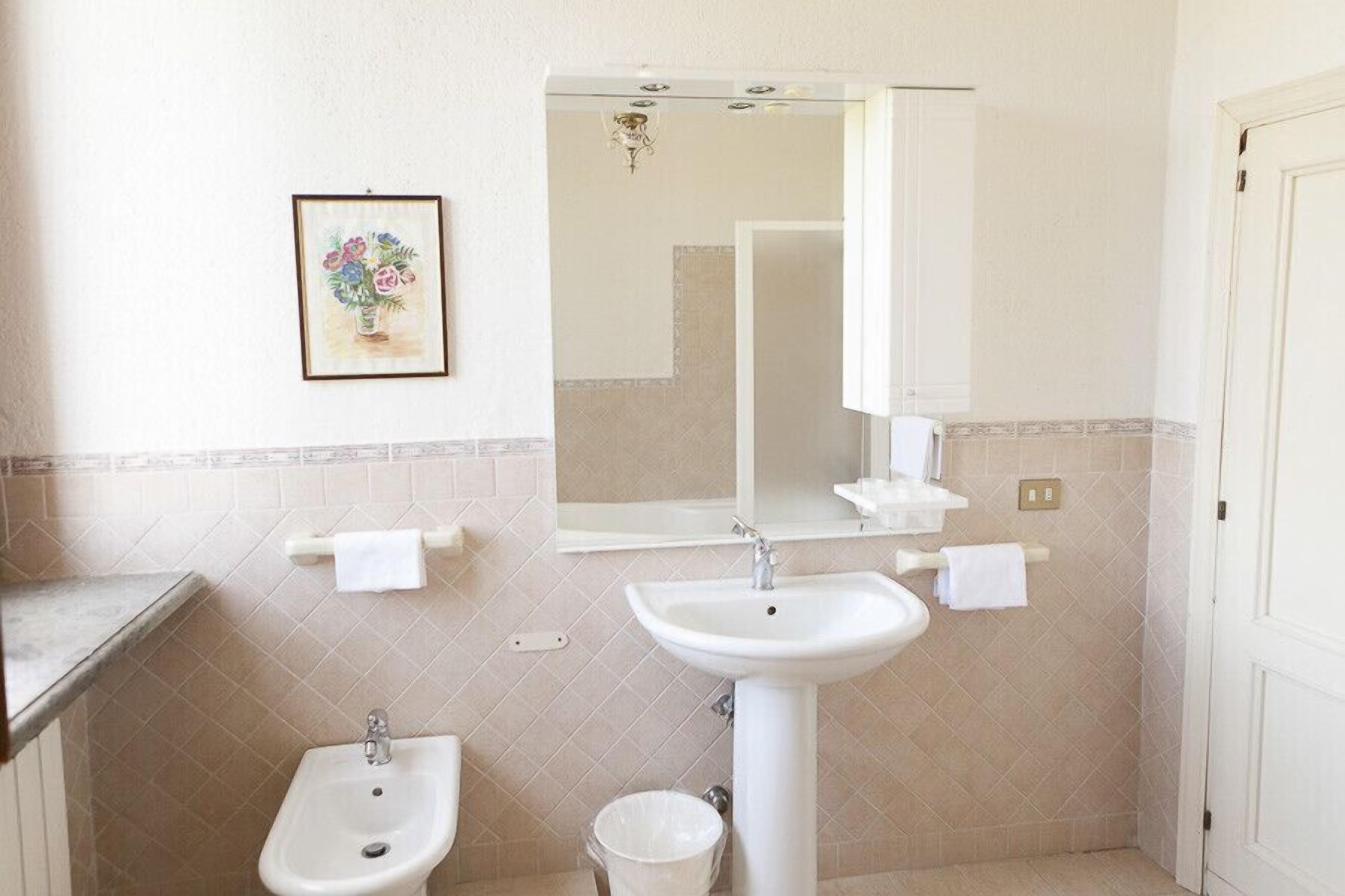 Une salle de bain avec un lavabo sur colonne, un bidet, un miroir et une œuvre d’art florale encadrée accrochée au mur.
