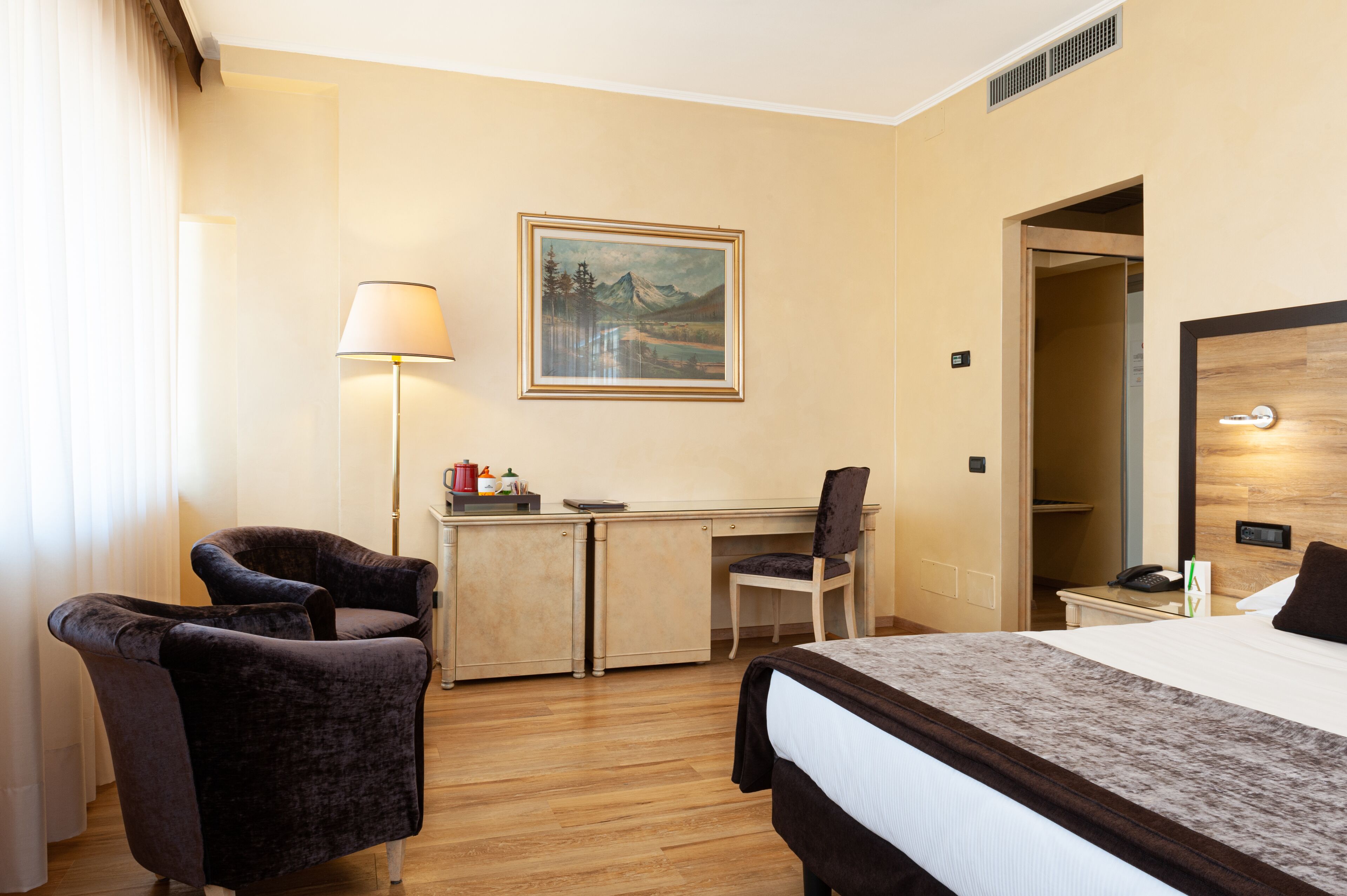 Una camera d'albergo con un letto, una scrivania, due sedie, una lampada e un quadro appeso al muro.