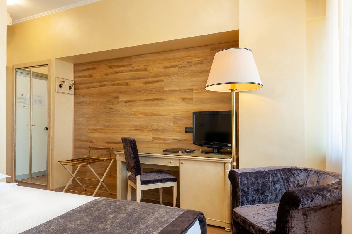 Een hotelkamer met een houten wand, een bureau met computer en televisie, een stoel, een bed en een lamp.