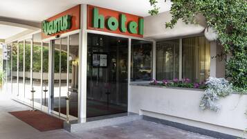 Entrada de hotel con puerta de cristal, un letrero rojo y verde, y un ventanal con flores.