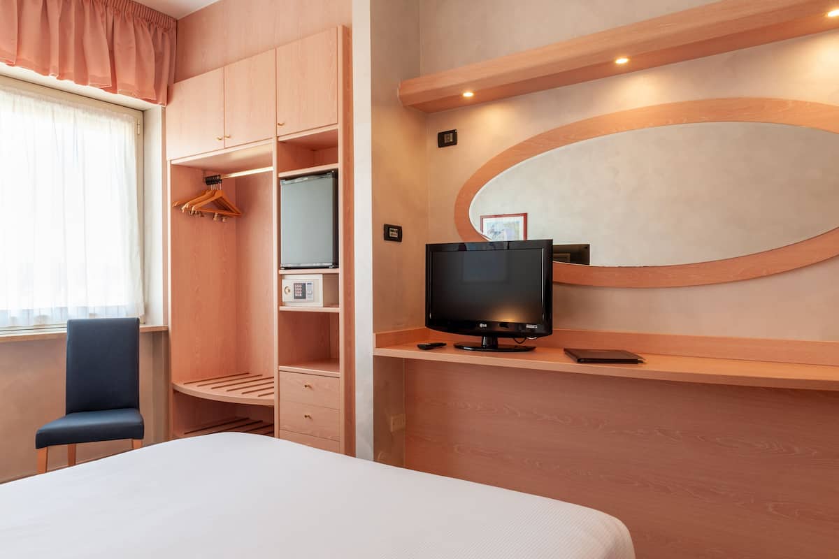 Een hotelkamer met een bed, een bureau met een computer, een televisie, een kledingkast en een raam met gordijnen.