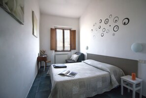 A bedroom with a bed, a bedside table, a chair, a radiator, and a window with curtains. - Il Ghiro (Florence)