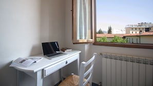 Une pièce avec un bureau blanc, un ordinateur portable et une chaise. Il y a une fenêtre donnant sur des immeubles et des arbres.