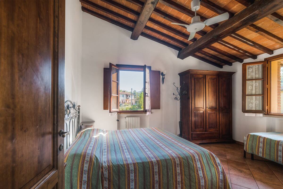Une chambre avec un plafond en bois, un lit recouvert d’une courtepointe à rayures, une armoire et une fenêtre donnant sur des immeubles.