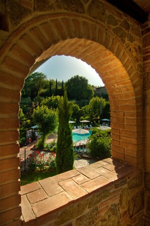 Une vue à travers une fenêtre en arc sur un espace piscine, avec un mur de briques, de la verdure et un ciel dégagé.